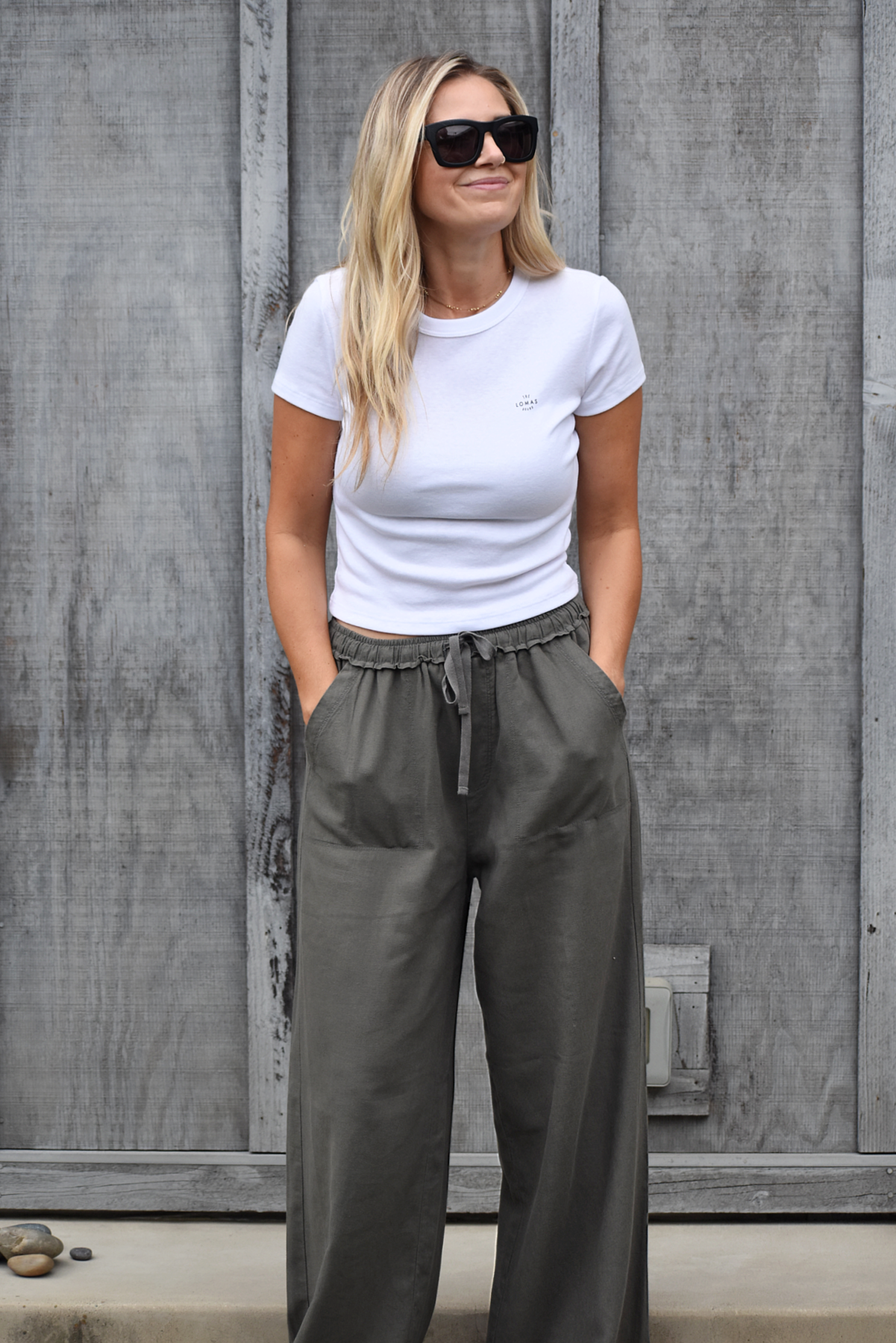 A woman standing in front of a weathered wooden fence. She's smiling and wearing a white t-shirt, olive pants, and dark sunglasses. Her long blonde hair falls past her shoulders.​​​​​​​​​​​​​​​​