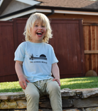 Toddler sitting on a stone curb laughing while wearing a soft teal blue t-shirt. Adorning the shirt is an iconic logo showcasing a saguaro with reaching limbs, an airborne bird, and an emerging sun, portrayed in a deep black shade. The phrase 'The Lomas Brand' is prominently displayed below the graphic.