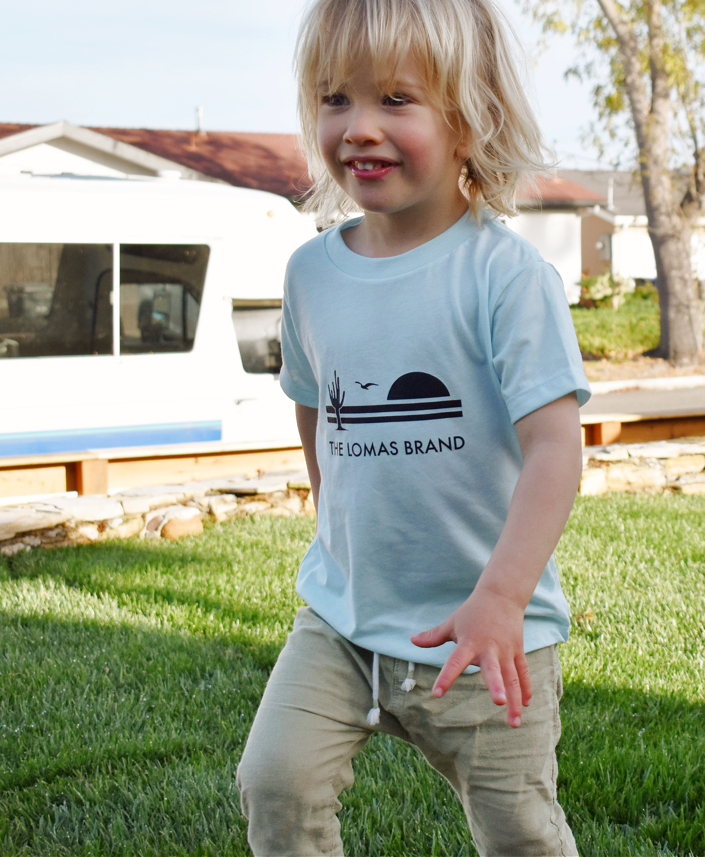 Toddler runs towards the camera with a smile while wearing a soft teal blue t-shirt. The shirt has The Lomas Brand logo with a saguaro cactus, bird, and a rising sun in black. 'The Lomas Brand' is printed below.