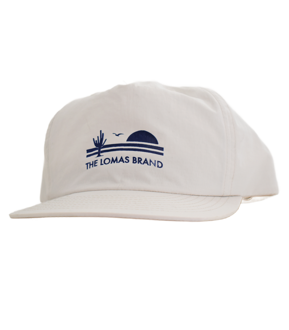 White baseball cap with a navy blue embroidered logo on the front. The logo shows a sunset scene with a cactus and "THE LOMAS BRAND" text. The cap is photographed straight-on against a white background.