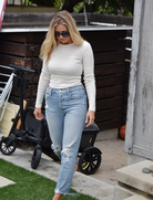 Blonde woman in sunglasses and white long-sleeved top walks on grass near a wagon or stroller. She's wearing ripped blue jeans.