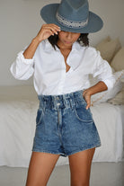 Woman with short black hair facing the camera wearing a white oxford shirt tucked into denim shorts. She has a blue felt boater-style hat and tips it forward with one hand. Her other hand is at her waist. A bed can be shown in the background.