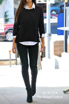 Mid body photo of a woman with long brown hair walking towards the camera. She has a black sweater over a white oxford shirt with the sleeves rolled up and the collar popping out, along with black pants, and black boots. The words "-WINTER-" and "The Abigail Oxford" are superimposed in the lower right of the photo.