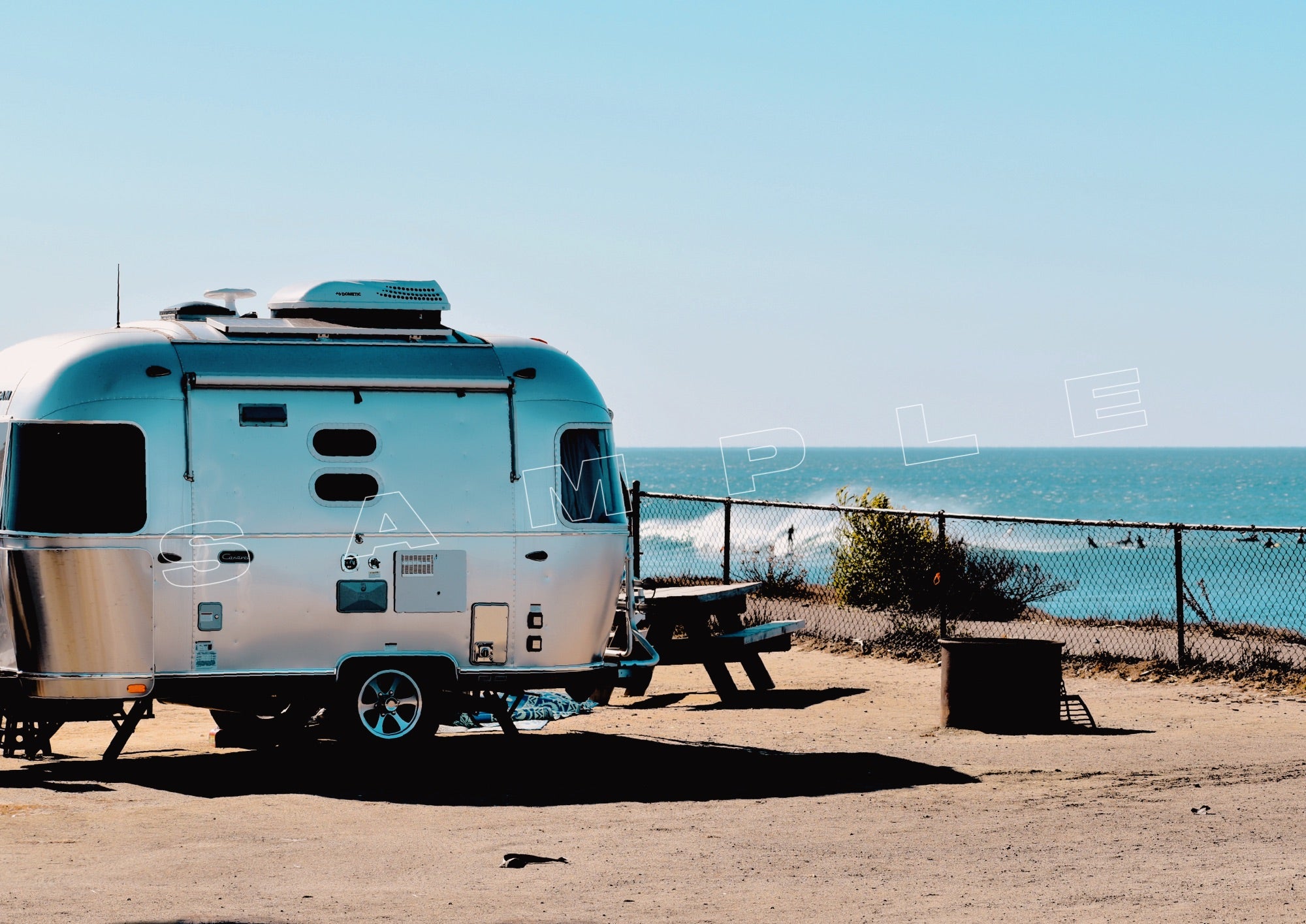 A silver Airstream trailer parked at a beach campsite with the ocean visible in the background. A picnic table and fence are nearby. The word "SAMPLE" is overlaid on the image.