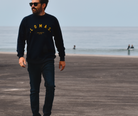 Man walking on a sandy beach wearing a navy sweatshirt with a yellow 'LOMAS' written in an arch, sunglasses, and jeans. The ocean is visible behind him, and there are some surfers in the water in the distance.