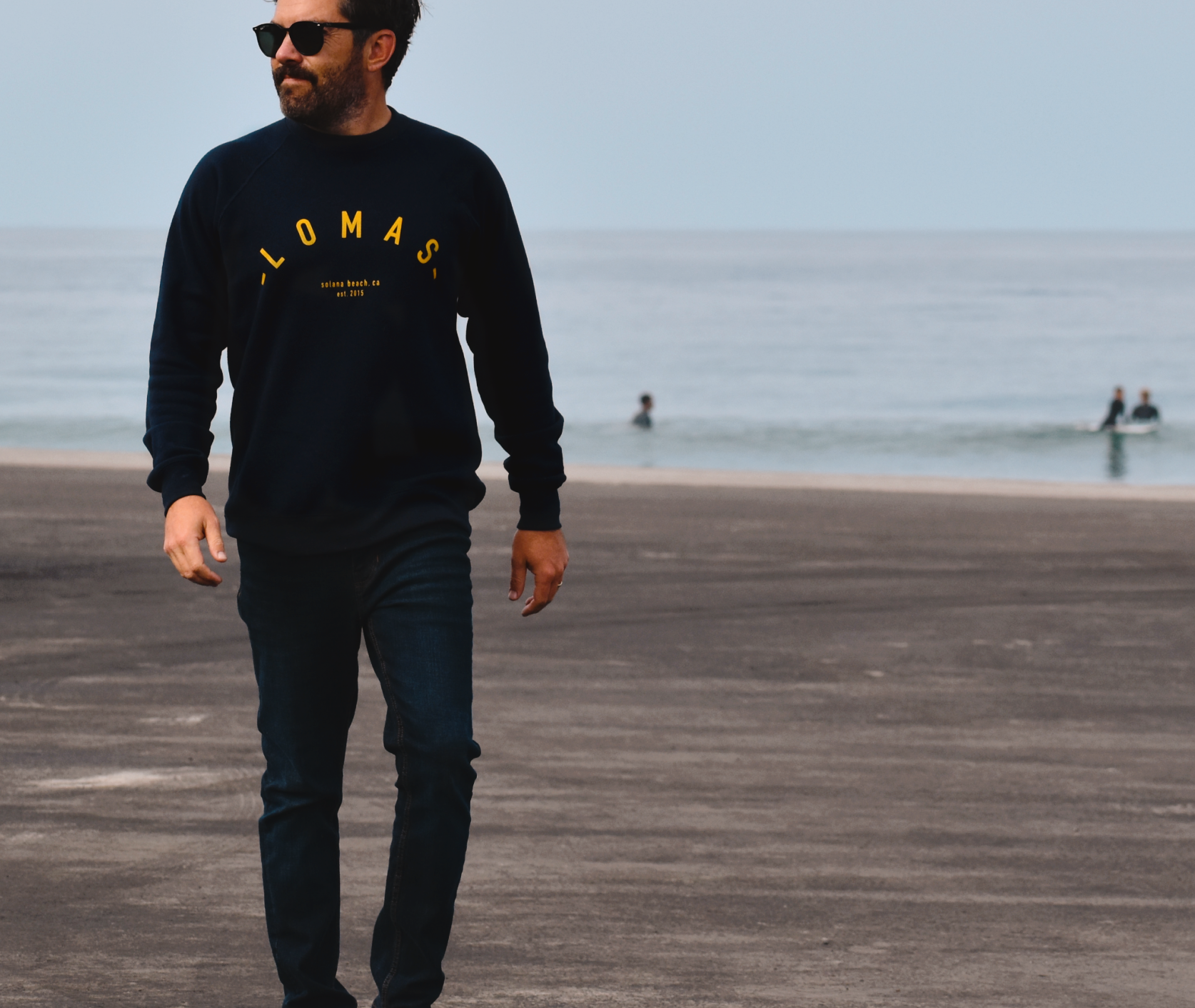 Man walking on a sandy beach wearing a navy sweatshirt with a yellow 'LOMAS' written in an arch, sunglasses, and jeans. The ocean is visible behind him, and there are some surfers in the water in the distance.