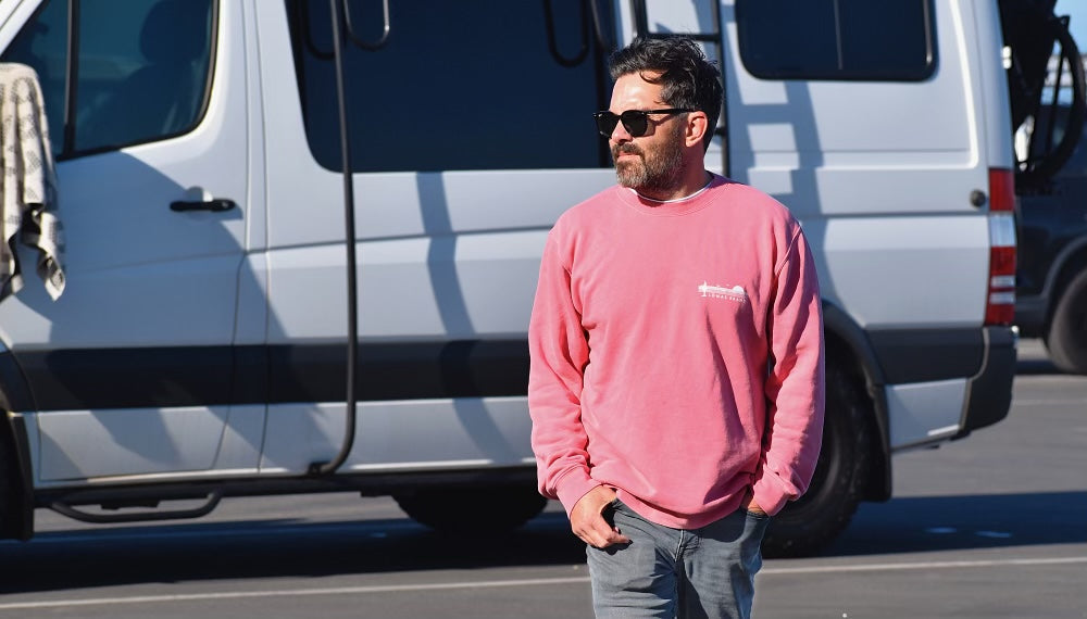 A man with a beard wearing sunglasses and a salmon colored sweatshirt standing in front of a white van. He has his hands in his pockets and is looking to the side.