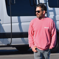A man with a beard wearing sunglasses and a salmon colored sweatshirt standing in front of a white van. He has his hands in his pockets and is looking to the side.
