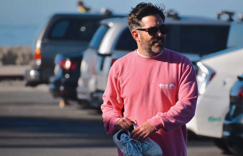 A man with a beard wearing sunglasses and a salmon colored sweatshirt. He is standing outdoors with vehicles visible in the background. Only his upper body is shown in the image.
