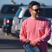 A man with a beard wearing sunglasses and a salmon colored sweatshirt. He is standing outdoors with vehicles visible in the background. Only his upper body is shown in the image.