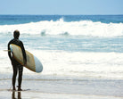 A surfer in a black wetsuit stands on the beach holding a surfboard, looking out at breaking waves. The word "SAMPLE" is superimposed on the image.