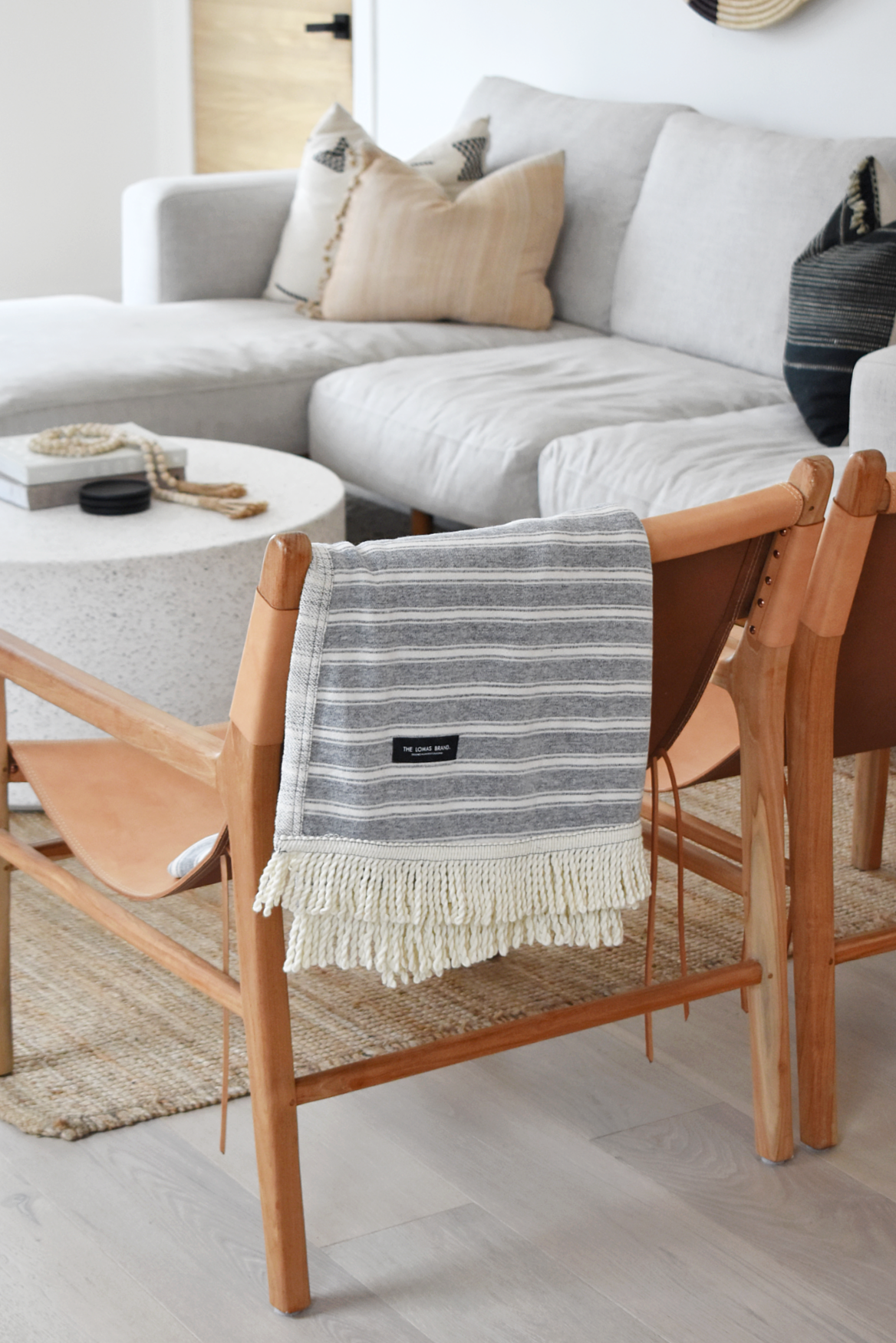 Grey and white striped blanket with fringe and a black patch that says "THE LOMAS BRAND," folded and hung on the back of a chair. A coffee table and a couch are in the background. The room looks very crisp and clean.