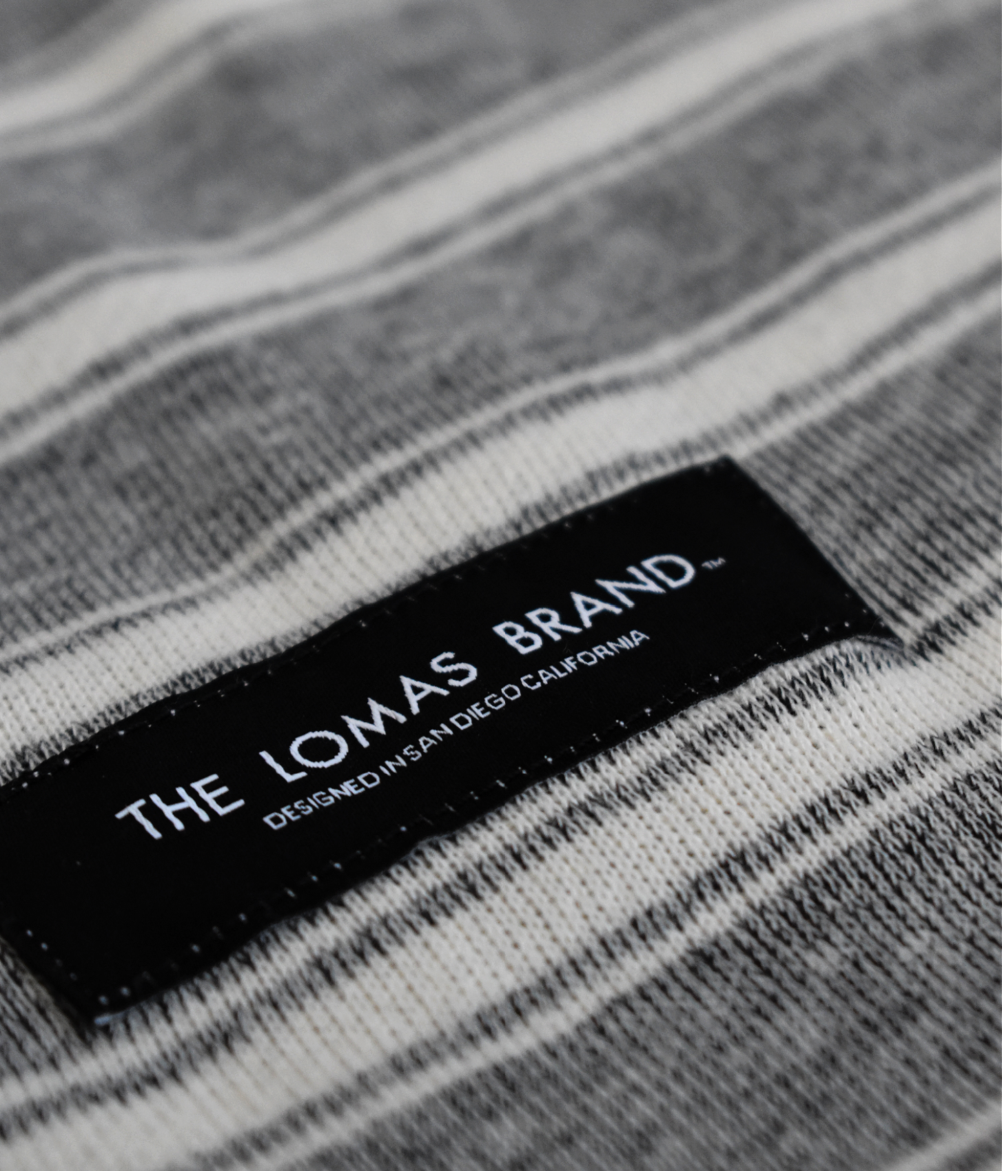 Close up of a grey and white blanket patch that says "THE LOMAS BRAND" with "DESIGNED IN SAN DIEGO CALIFORNIA" underneath.