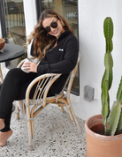 A woman sitting in a wicker chair on a porch or patio. She's wearing sunglasses, a black quarter zip sweater with a small white logo, and holding a mug. There's a cactus plant next to her.