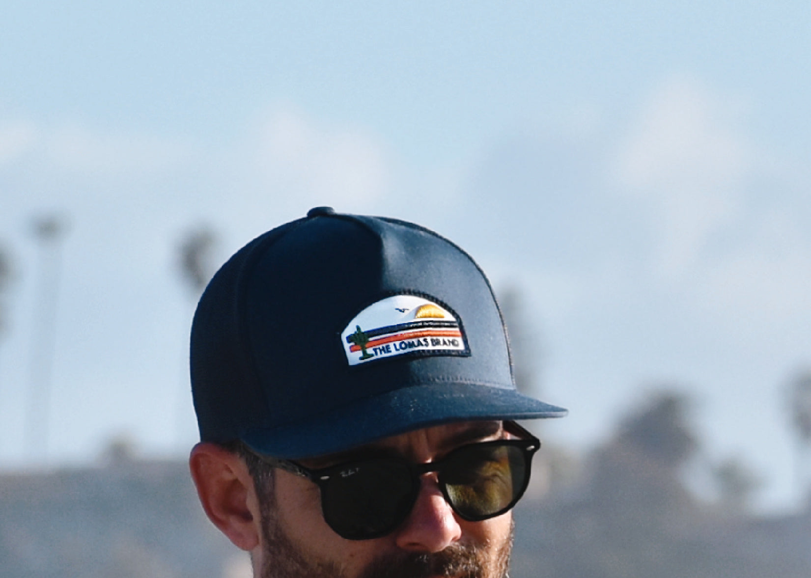 Close-up of a navy blue baseball cap with a logo patch showing a sunset scene and the text "THE LOMAS BRAND".