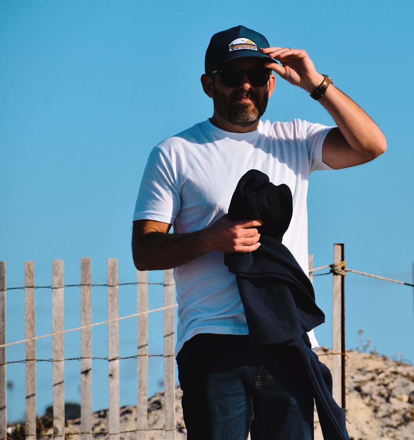 A man wearing a white t-shirt and navy cap with a logo, adjusting his hat while standing in front of a wooden fence on a sunny day. The logo on the hat says "THE LOMAS BRAND" and has a scene with a cactus, a sun, and a bird.