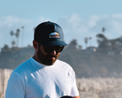 A man in a white t-shirt and navy cap with a logo, looking down while standing in front of a blurred background of palm trees and hills. The logo on the hat says "THE LOMAS BRAND" and has a scene with a cactus, a sun, and a bird. 