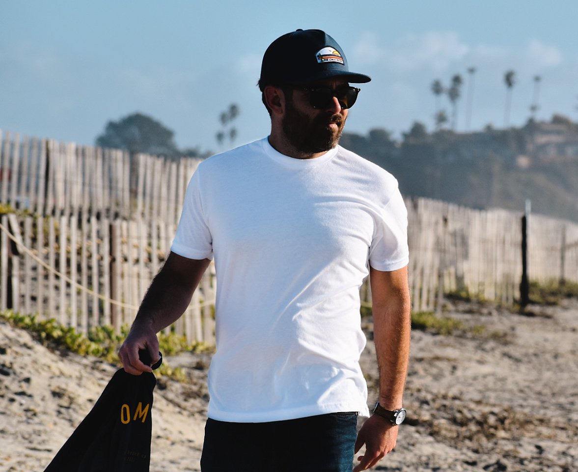 A man wearing a white t-shirt, navy cap with a logo, and sunglasses standing on a beach with a wooden fence in the background. The logo on the hat says "THE LOMAS BRAND" and has a scene with a cactus, a sun, and a bird. He is holding a navy sweatshirt.