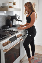 Woman with wavy dark blonde hair stands in a kitchen near an espresso machine. She smiles and looks at her coffee mug. She is wearing black joggers and a black sports bra. Both the joggers and the sports bra have a small white logo printed on them, featuring a saguaro cactus, a flying bird, and a rising sun. 