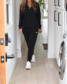 Mid body photo of a woman wearing black joggers and a black quarter zip sweater. Both articles of clothing have a small white logo, courtesy of The Lomas Brand. She is standing in a clean white hallway and a washing machine is visible.