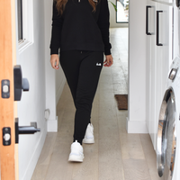 Mid body photo of a woman wearing black joggers and a black quarter zip sweater. Both articles of clothing have a small white logo, courtesy of The Lomas Brand. She is standing in a clean white hallway and a washing machine is visible.