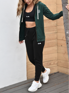 Woman with wavy hair wearing a forest green zip up sweatshirt with a black sports bra underneath. She also has black joggers and is wearing white shoes. Everything but the shoes has a white icon, stylized with a cactus, a bird, and a rising sun. She is smiling and looking down while holding on to a tree trunk with one arm.  