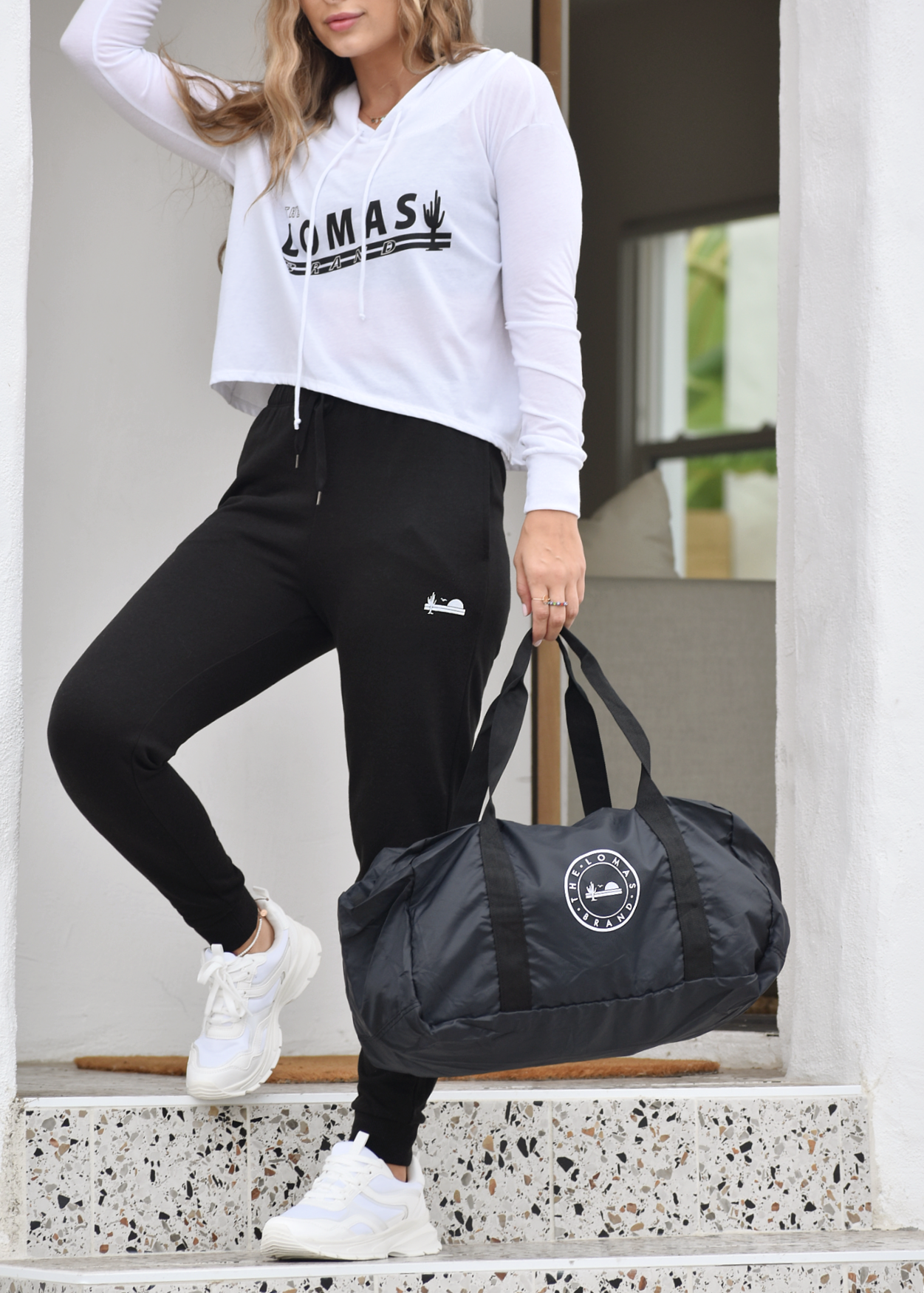 A woman in a white cropped hoodie with "LOMAS" printed in black. She's wearing black leggings and white sneakers, holding a black duffel bag. The outfit appears to be athletic wear.