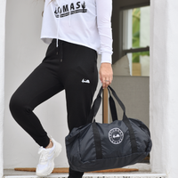 A woman in a white cropped hoodie with "LOMAS" printed in black. She's wearing black leggings and white sneakers, holding a black duffel bag. The outfit appears to be athletic wear.