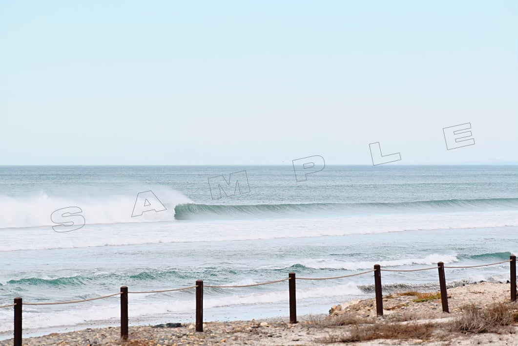 A breaking ocean wave with white foam visible. A rope fence and sandy beach are in the foreground. The sky is clear and the word "SAMPLE" is overlaid on the image. 