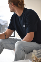 A close-up of a man's head and upper back, showing his hair tied in a bun and the back of his navy t-shirt with a small green, orange, and white logo patch visible on the upper right chest.