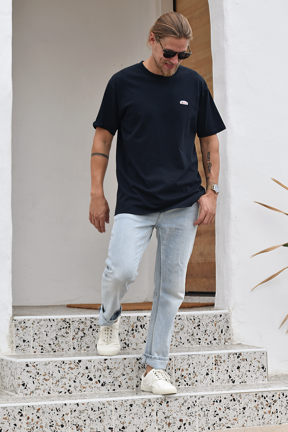 A man standing on what appears to be terrazzo steps, wearing a navy t-shirt and light blue jeans. The shirt has a small green, orange, and white logo patch visible on the upper right chest. He's looking down and smiling.