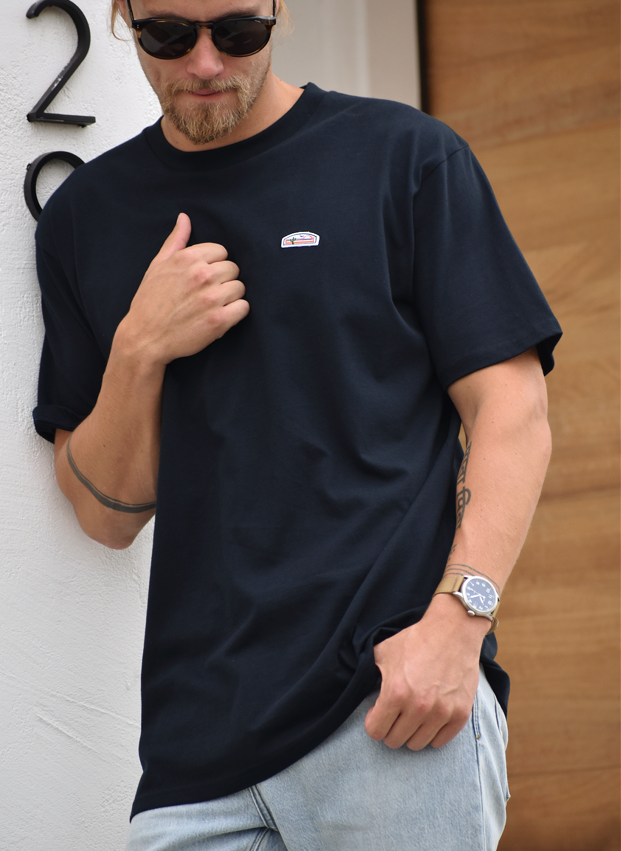 A close view of a man's upper body, showing his beard, sunglasses, and a navy t-shirt. The shirt has a small green, orange, and white logo patch visible on the upper right chest. The man is making a gesture with his hand near his chest.