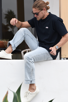 A man sitting on a ledge or bench, wearing a navy blue t-shirt with a small orange, green, and white logo patch on the chest, light blue jeans, and white sneakers. He has long blonde hair tied back and is wearing sunglasses.