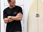 Man wearing a black t-shirt with a small white logo on the chest. The logo says "THE LOMAS BRAND" in a circular font around a saguaro cactus, a bird, and a rising sun. He is standing next to a surfboard, has blonde hair and a beard, and is smiling slightly while looking to the side.