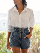 Mid body photo of a woman with short dark hair wearing a white button down oxford shirt tucked into high waisted short jeans. Her hand is in her pocket and she looks off camera. 